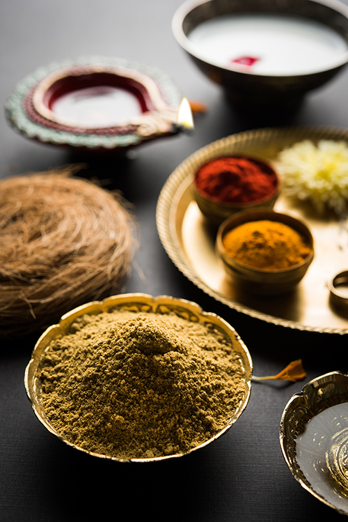 Brass bowls filled with colorful powdered spices and herbs, arranged on a dark surface with decorative plates and a lit oil lamp in the background, evoke a festive ambiance inspired by holistic care and integrative medicine.