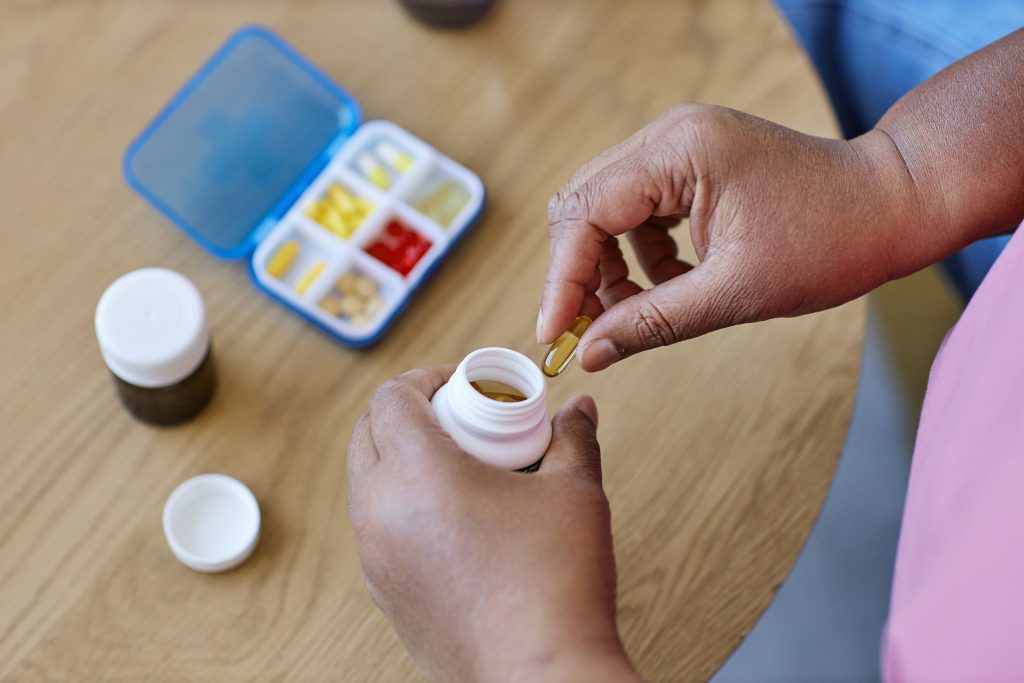 A person holds a yellow pill above an open medicine bottle, reflecting a mindful approach to wellness. On the table are two medicine bottles and a blue pill organizer with various pills sorted into compartments.