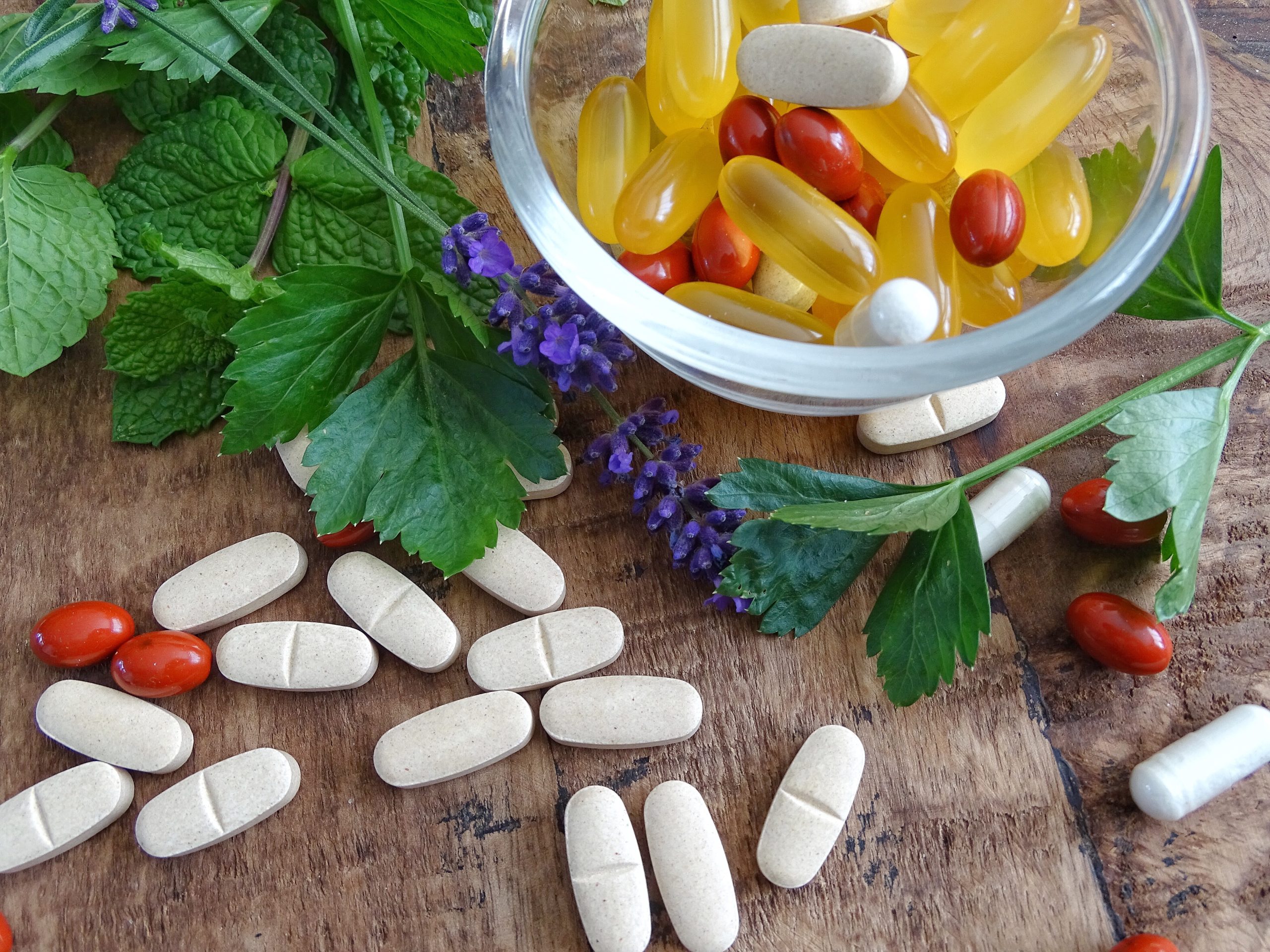 A glass bowl with yellow and red capsules and white tablets sits on a wooden surface, surrounded by scattered pills and fresh green herbs with purple flowers, reflecting the balance of wellness and integrative medicine.