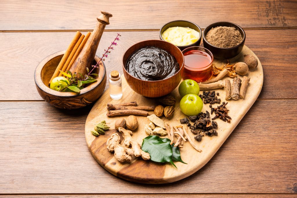 A wooden board displays various Ayurvedic herbs, roots, amla fruits, spices, powders, and pastes—suggesting a traditional herbal preparation that celebrates wellness and integrative medicine.
