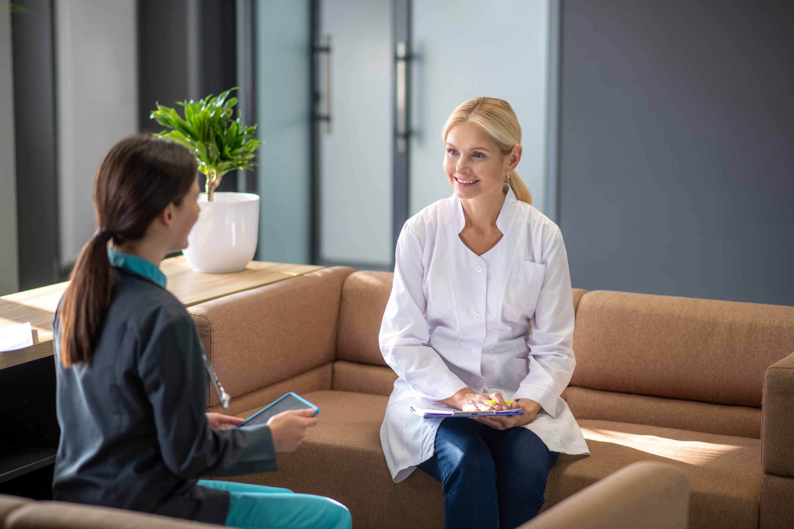 Two women sit on a tan couch in a modern office. One, in a white lab coat and holding a clipboard, smiles while discussing integrative medicine with the other, who holds a tablet. A potted plant rests on the table beside them.