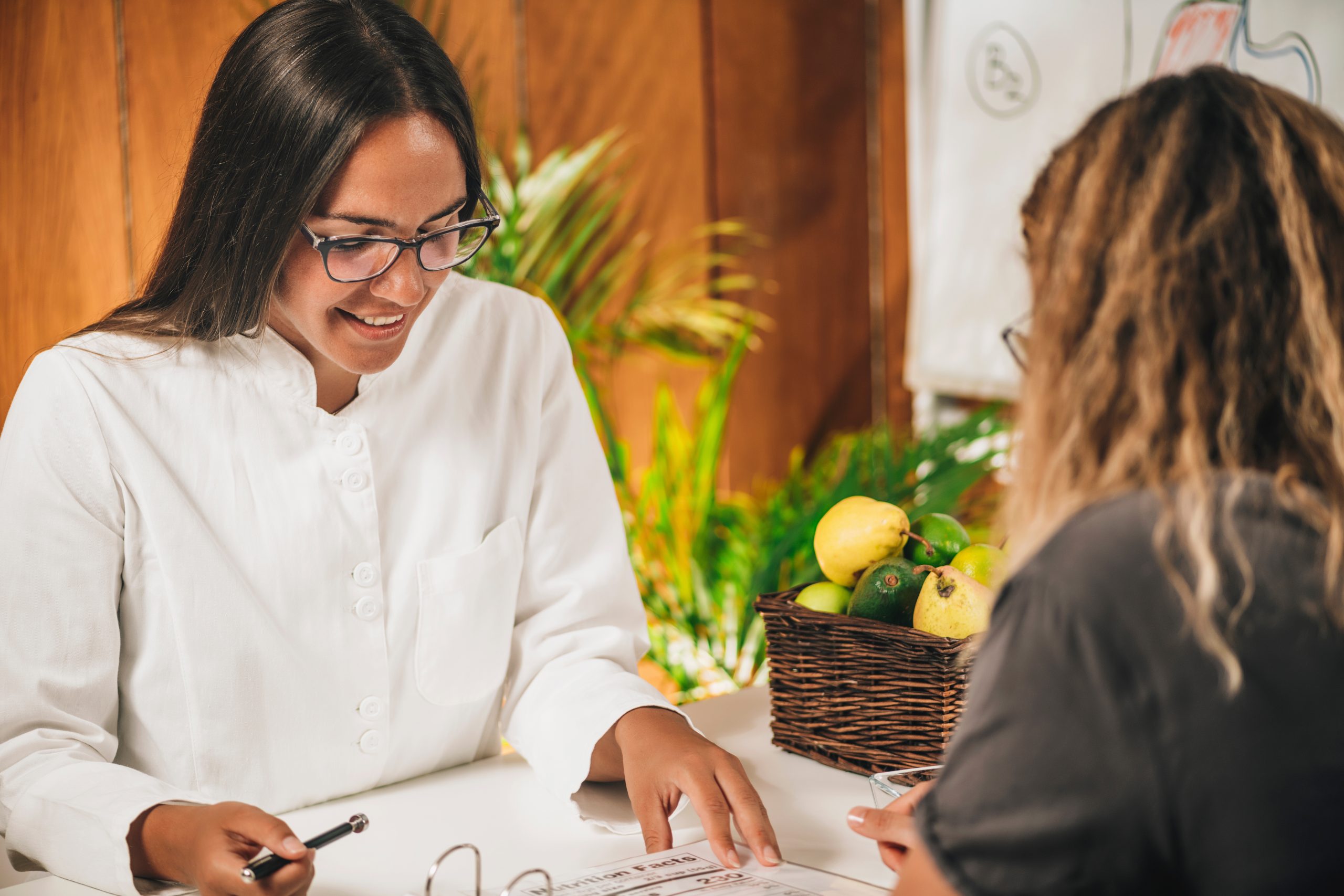 A woman sits at a table while another provides nutritional guidance, writing on a paper as part of a holistic care approach to wellness.