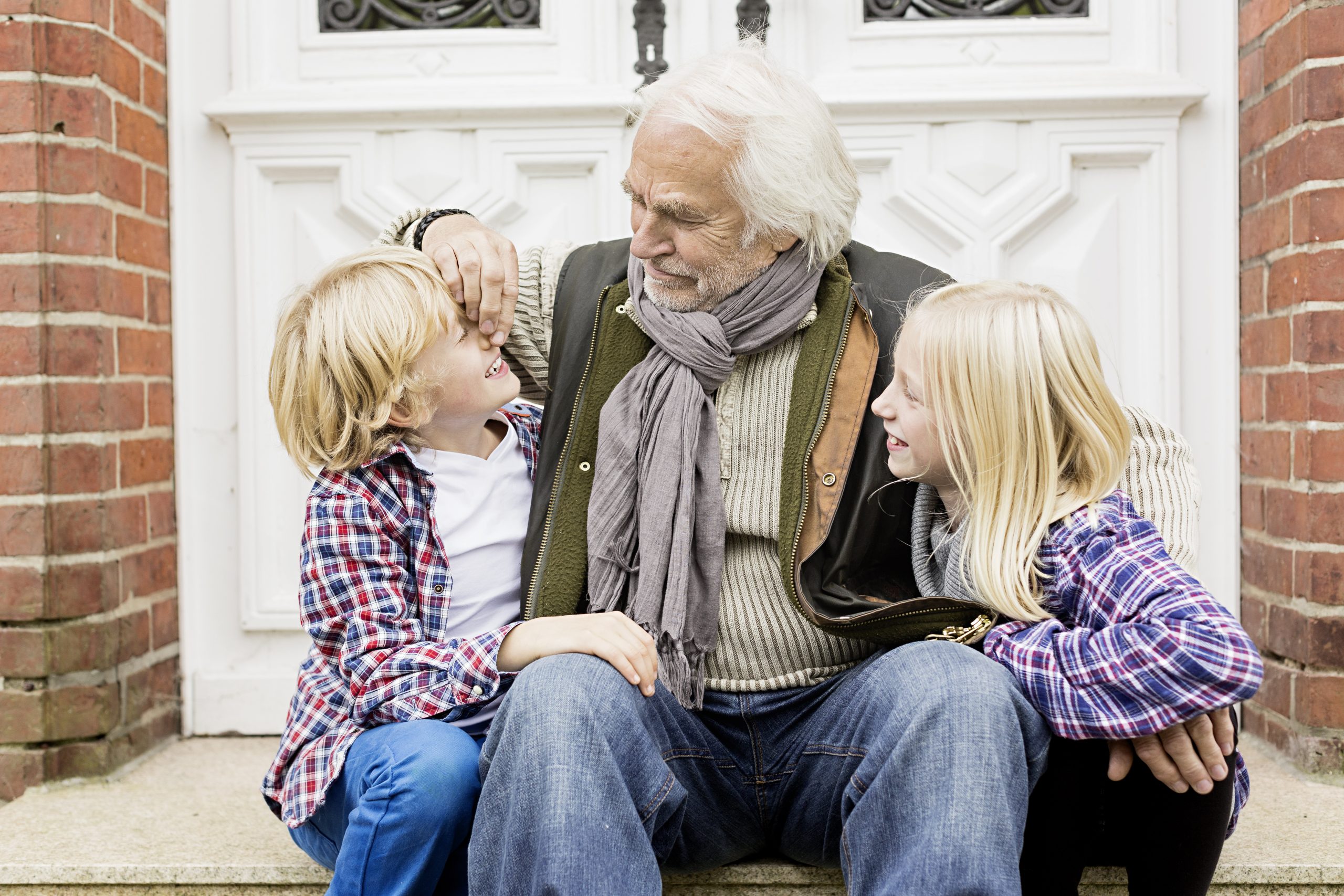 An elderly man sits on steps outside with a young boy and girl, smiling warmly, embodying the spirit of holistic care. The children, both blonde and wearing plaid shirts, look up at him affectionately. The background shows a white door and red brick walls.