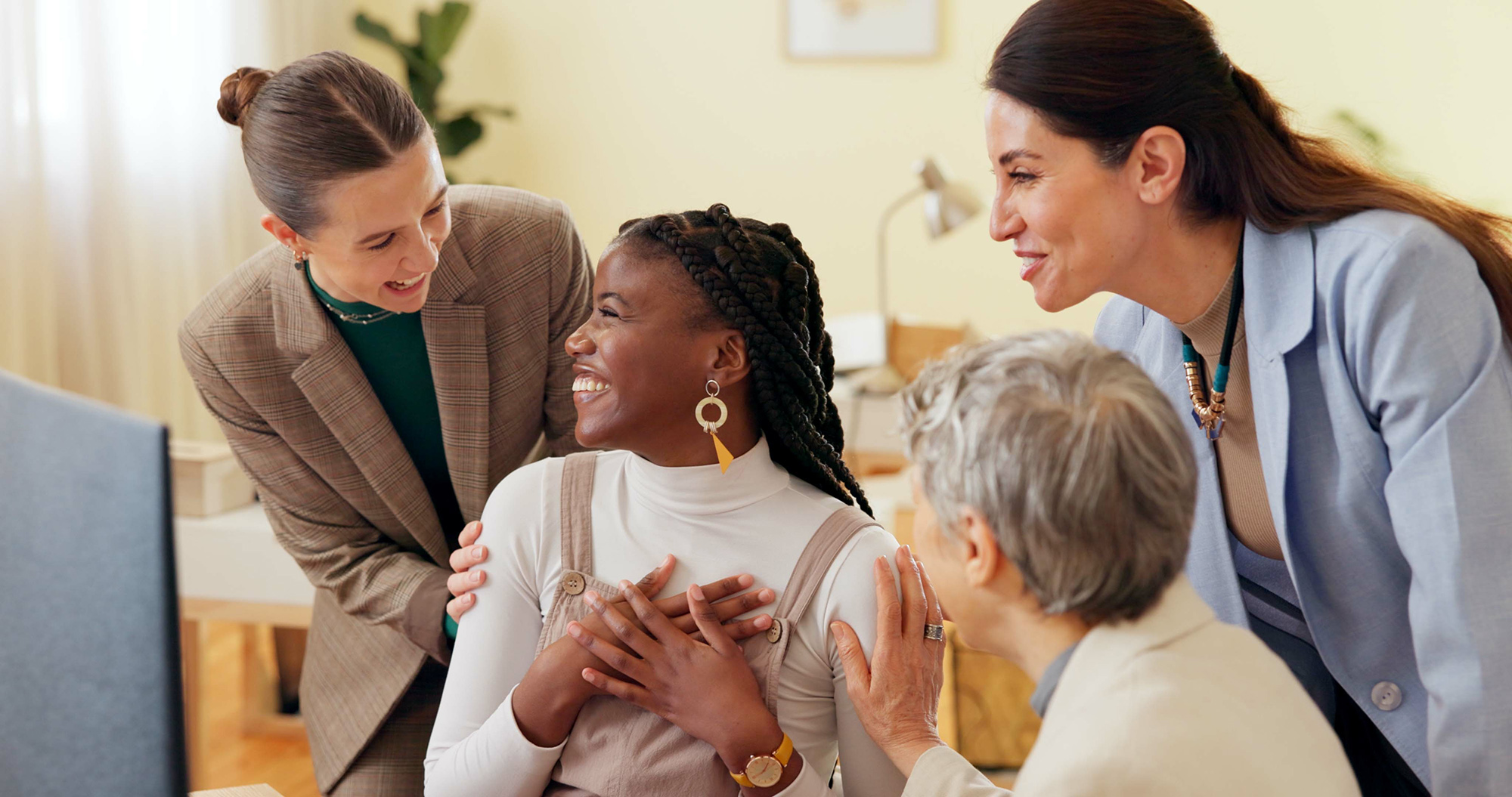 Four women, dressed in business attire, gather around a seated colleague who smiles joyfully with her hands on her chest. The others smile and touch her shoulders in a supportive, celebratory office setting reflecting holistic care values.