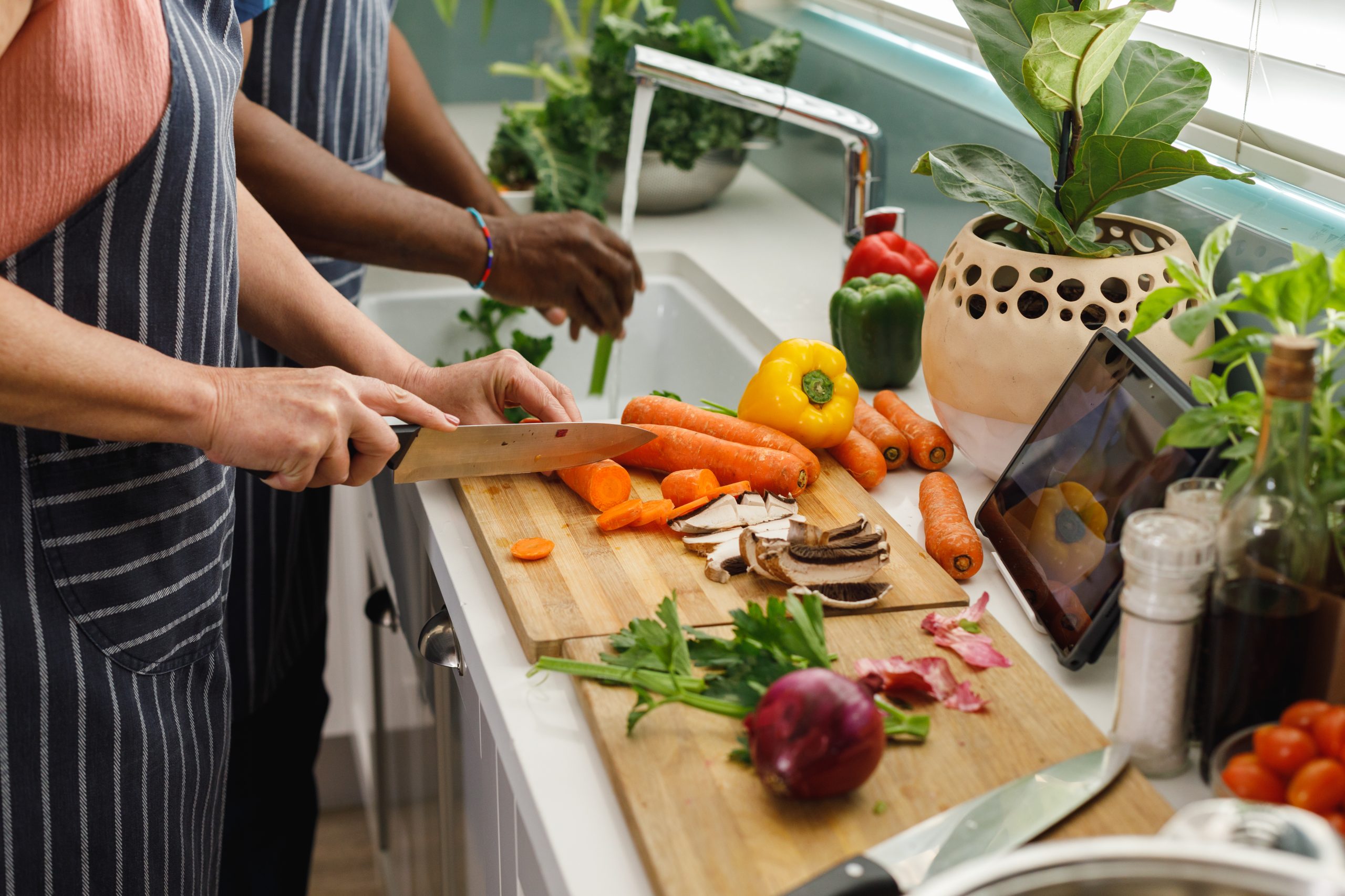 Two people preparing vegetables together in a kitchen, embracing holistic care as they slice carrots and mushrooms on a wooden cutting board; fresh produce surrounds them, while a tablet—perhaps with Heidelberger Integrative wellness tips—rests by the sink.