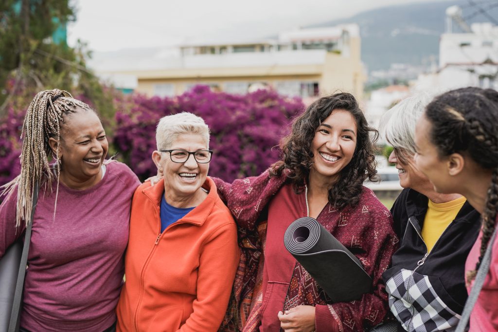 Five women of different ages stand outdoors in York PA, smiling and laughing together. One holds a yoga mat, reflecting the spirit of integrative medicine. They wear casual clothes, with purple flowers and buildings in the background.