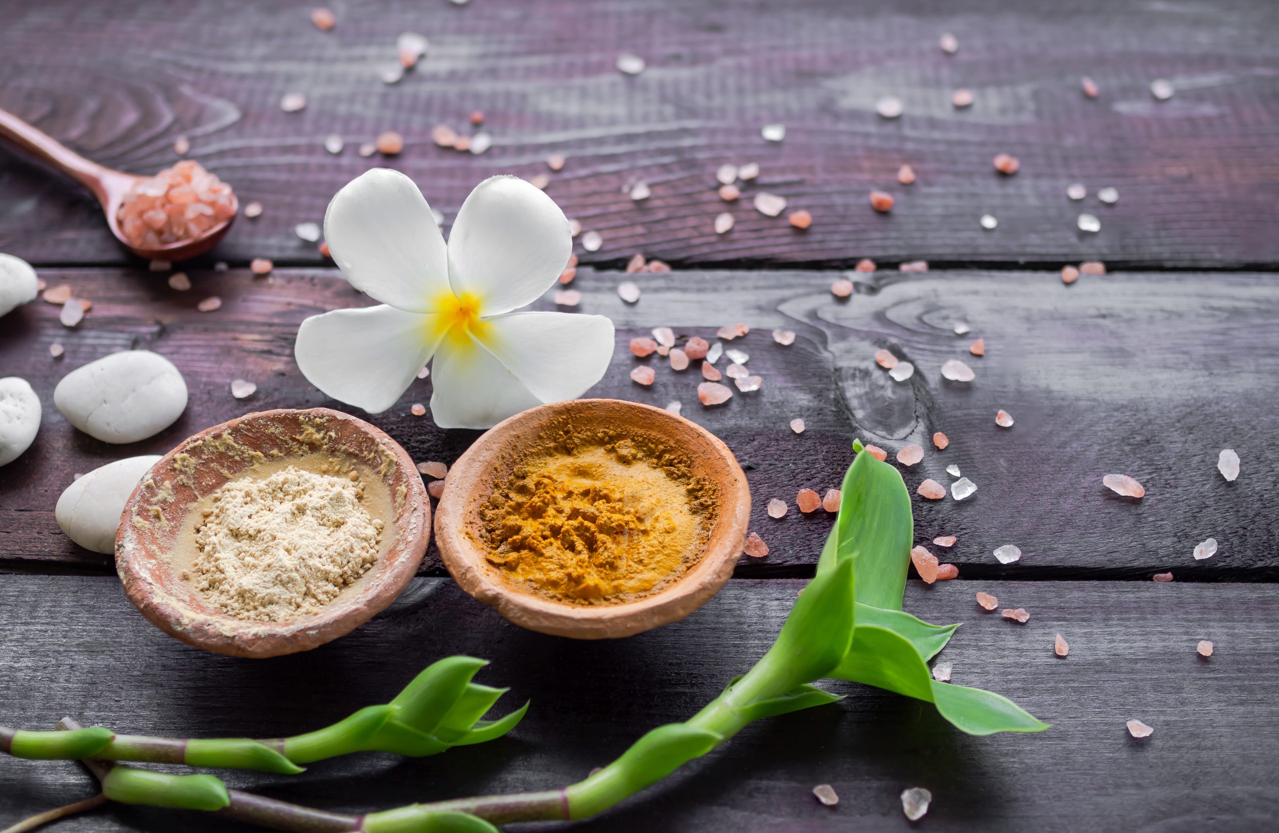 Two small wooden bowls with powdered spices, a white flower, green leaves, pink Himalayan salt, and white stones are arranged on a rustic wooden surface, evoking a natural wellness atmosphere inspired by Heidelberger Integrative in York PA.