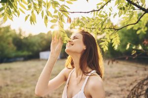 A young woman with long hair smiles and gently touches green leaves on a tree branch in a sunlit outdoor setting, embodying the spirit of wellness promoted by Heidelberger Integrative, while wearing a white sleeveless top.