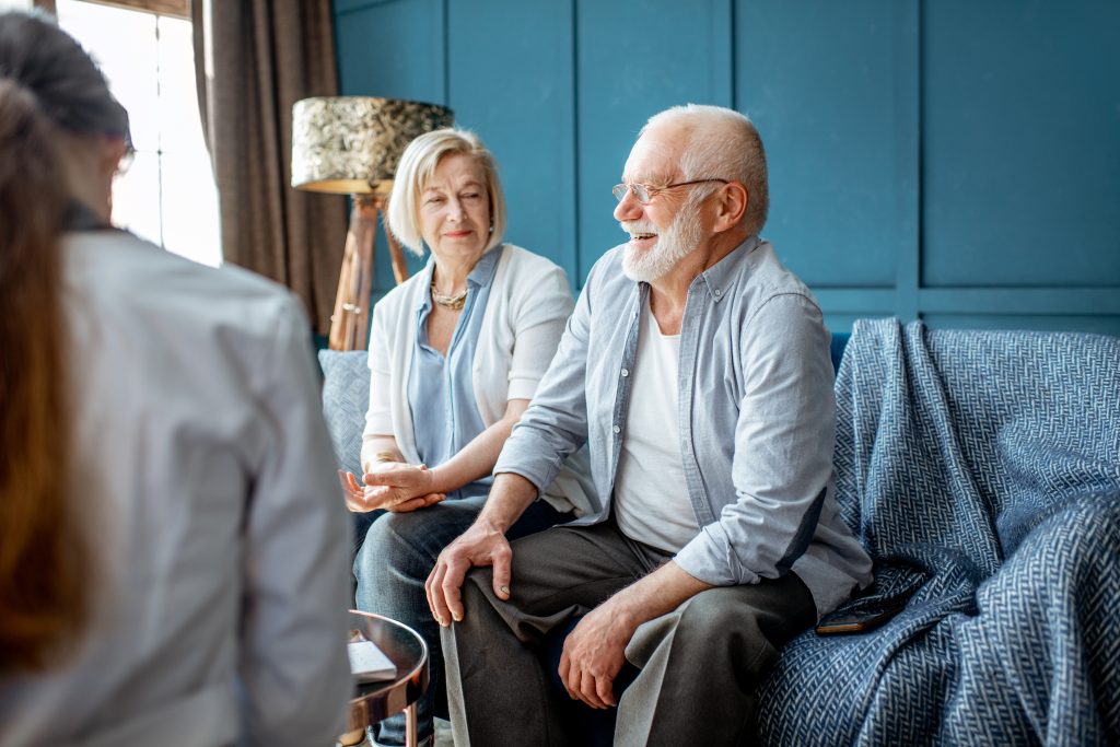 An elderly couple sits on a couch, smiling and talking to a woman in a white coat, possibly a healthcare professional, discussing holistic care in their cozy living room with blue walls.