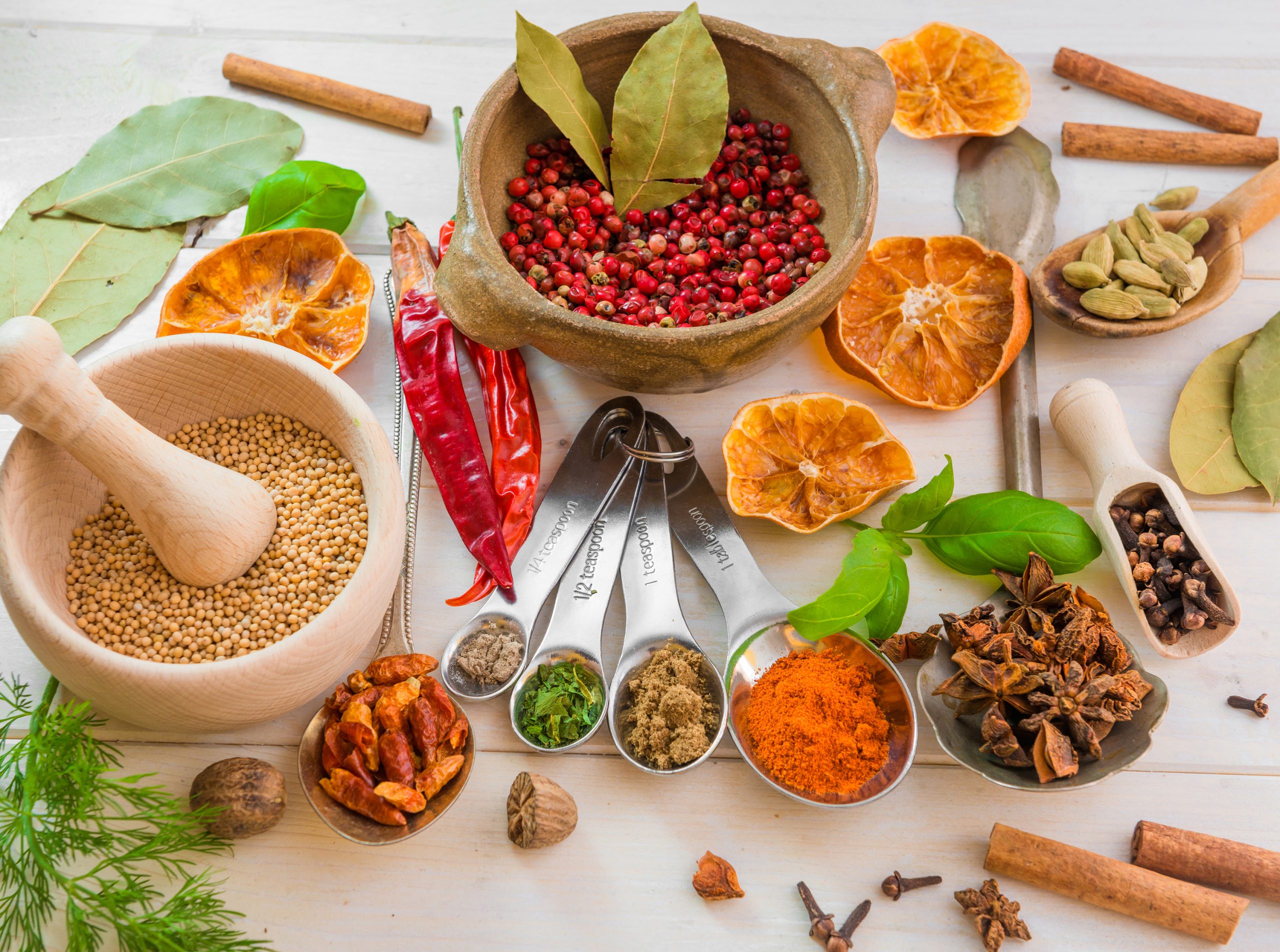 Assorted spices and herbs, often used in integrative medicine for nutritional guidance and holistic care, including mustard seeds, red chili peppers, dried orange slices, bay leaves, cardamom, cinnamon sticks, star anise, and ground spices on a white surface.