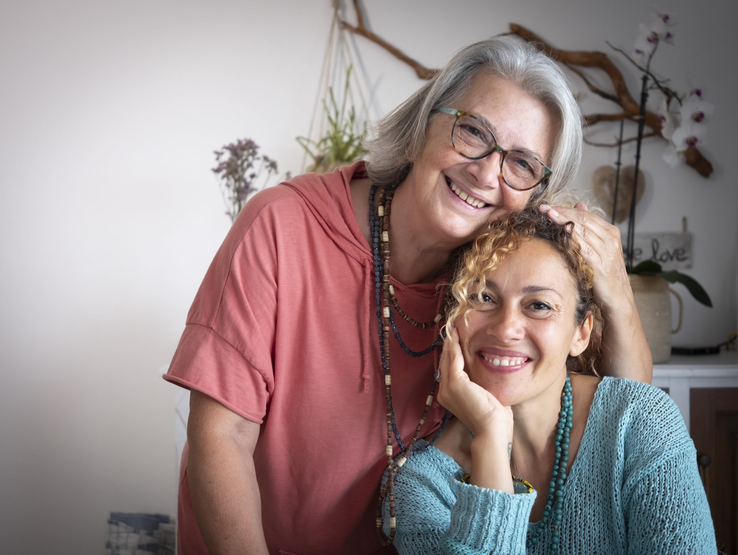 Two women smiling warmly at the camera, embodying the joy that comes from holistic care—one with gray hair and glasses embracing the other with curly hair, both appearing happy and relaxed in a cozy indoor setting.
