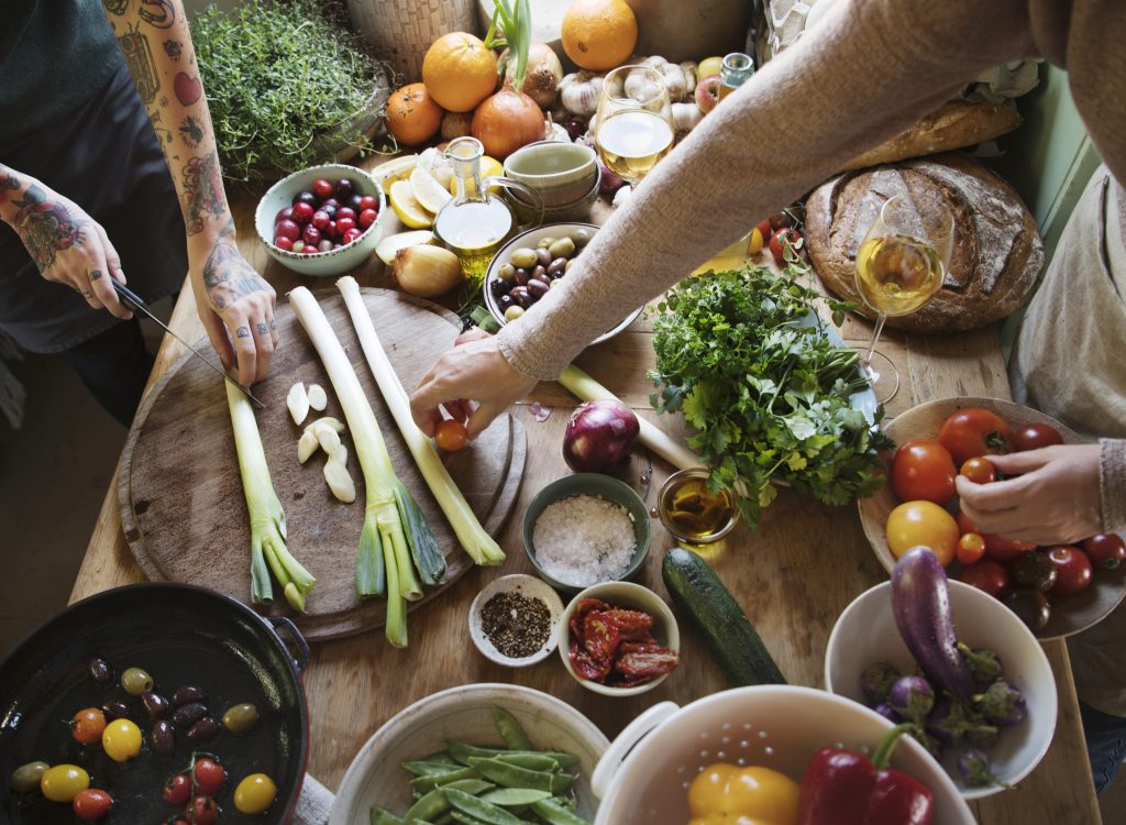 Three people prepare fresh vegetables and fruits on a wooden table filled with tomatoes, leeks, herbs, olives, and spices, creating a vibrant scene that reflects holistic care and a shared passion for wellness.