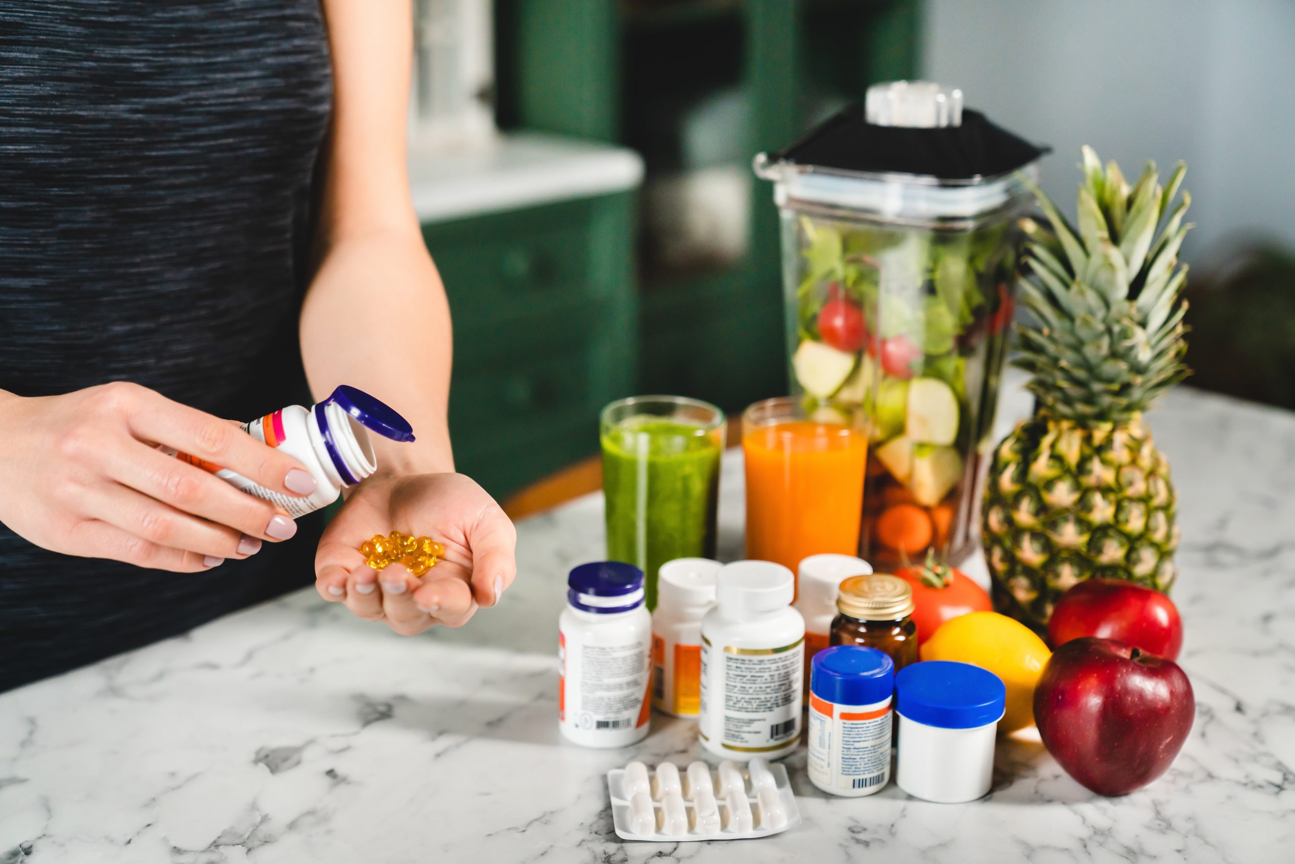 A person pours supplements into their hand next to pill bottles, a blender filled with fruit and vegetables, glasses of juice, and fresh produce—embracing a Heidelberger Integrative approach to wellness through integrative medicine and holistic care.
