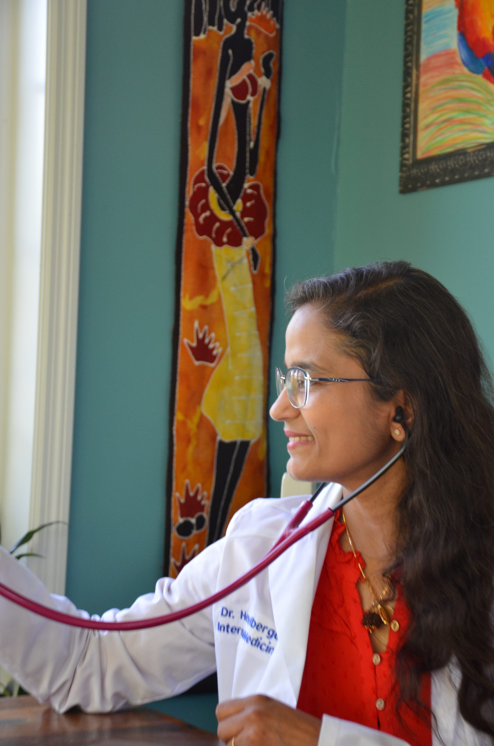 A doctor in a white coat and red blouse uses a stethoscope, smiling while seated indoors. Sunlight streams through the window, illuminating colorful artwork on the turquoise wall—reflecting her passion for integrative medicine and wellness in York PA.