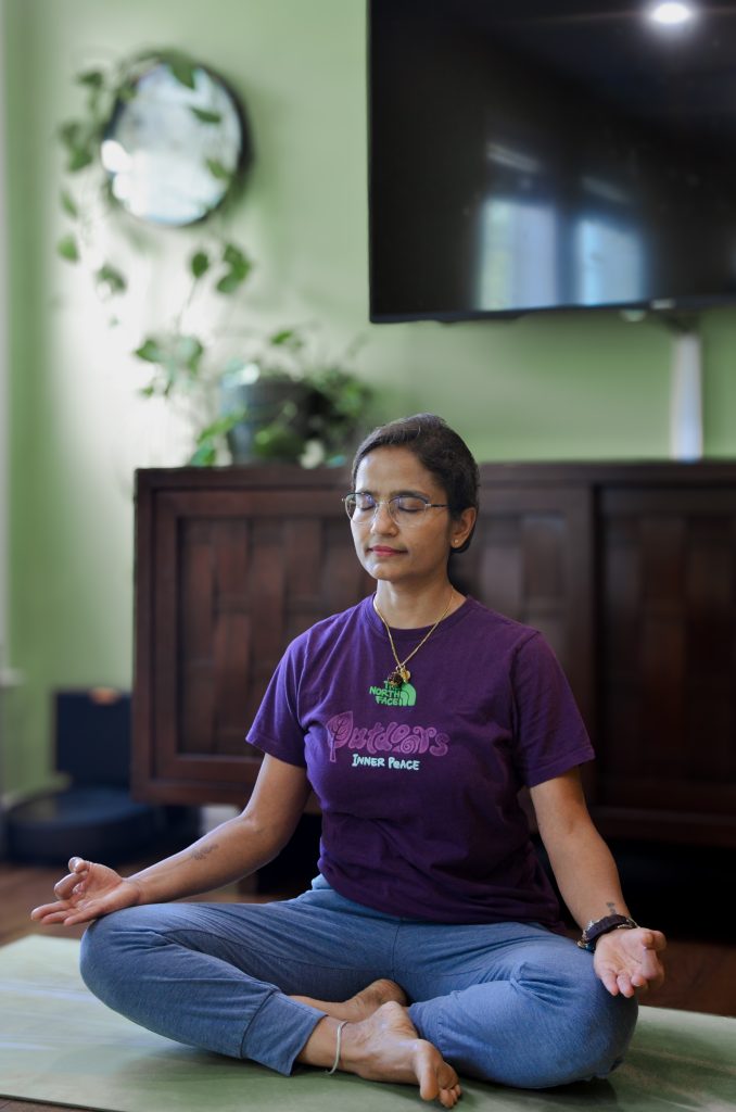 A woman with short hair and glasses sits cross-legged on the floor, meditating with eyes closed. She wears a purple t-shirt and blue pants. The serene setting reflects the calming atmosphere of integrative medicine at Heidelberger Integrative in York PA.