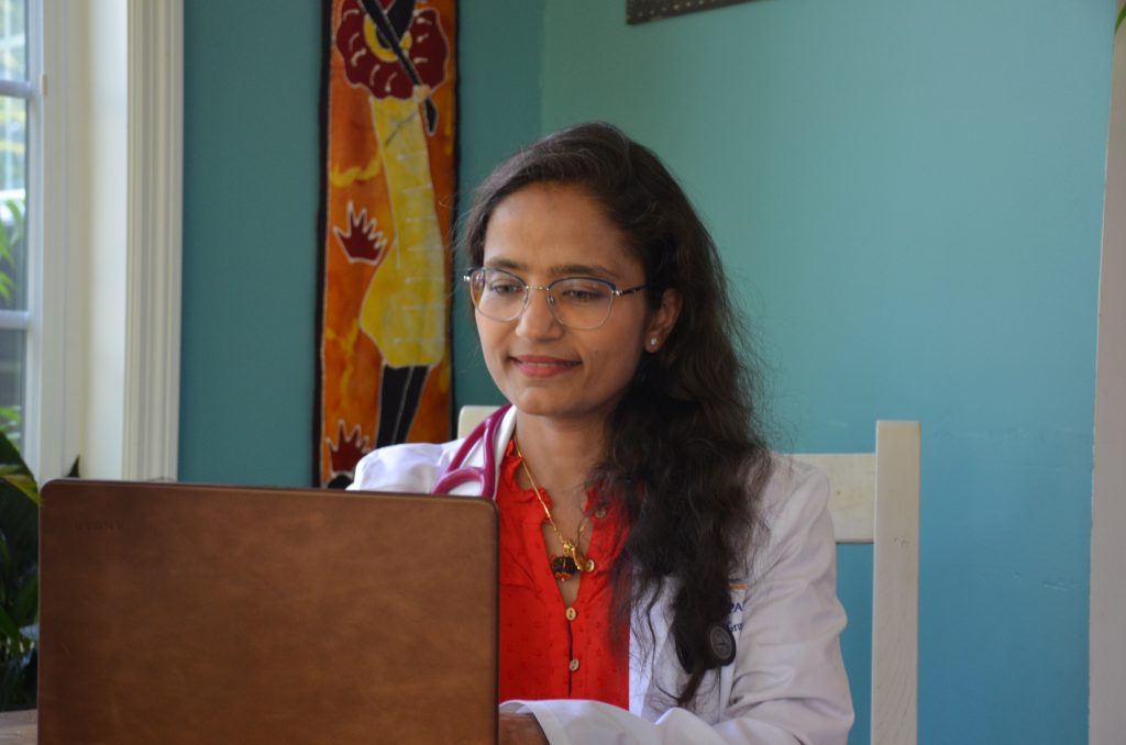 A woman wearing a white lab coat and stethoscope sits at a desk in York PA, looking at a laptop with a slight smile. She has long, wavy hair and eyeglasses. The background features colorful wall art and a blue wall, reflecting integrative medicine wellness.