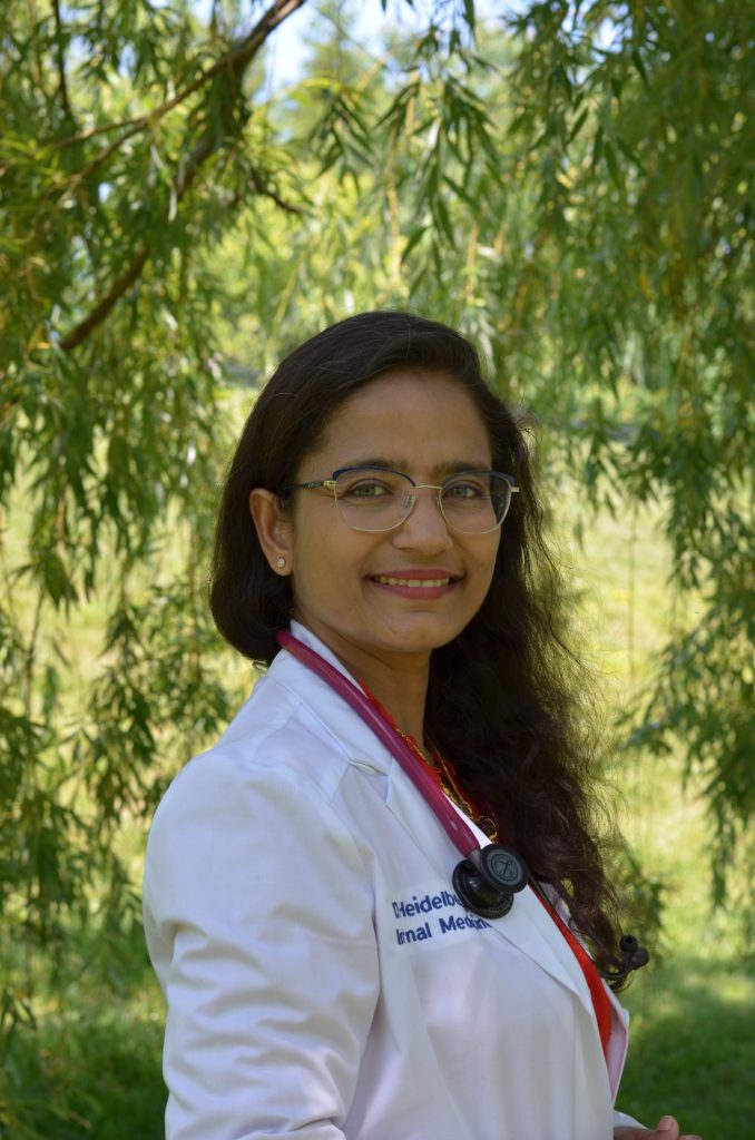 A woman in a white medical coat with a stethoscope around her neck smiles outdoors in front of leafy green trees, embodying holistic care and wellness.