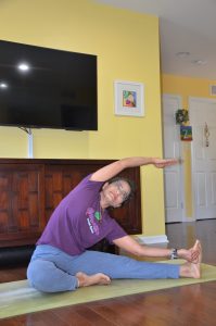 A woman in glasses and a purple shirt practices holistic care on a yoga mat indoors, stretching sideways near a TV and wooden cabinet in a bright room—embracing wellness inspired by Heidelberger Integrative principles.