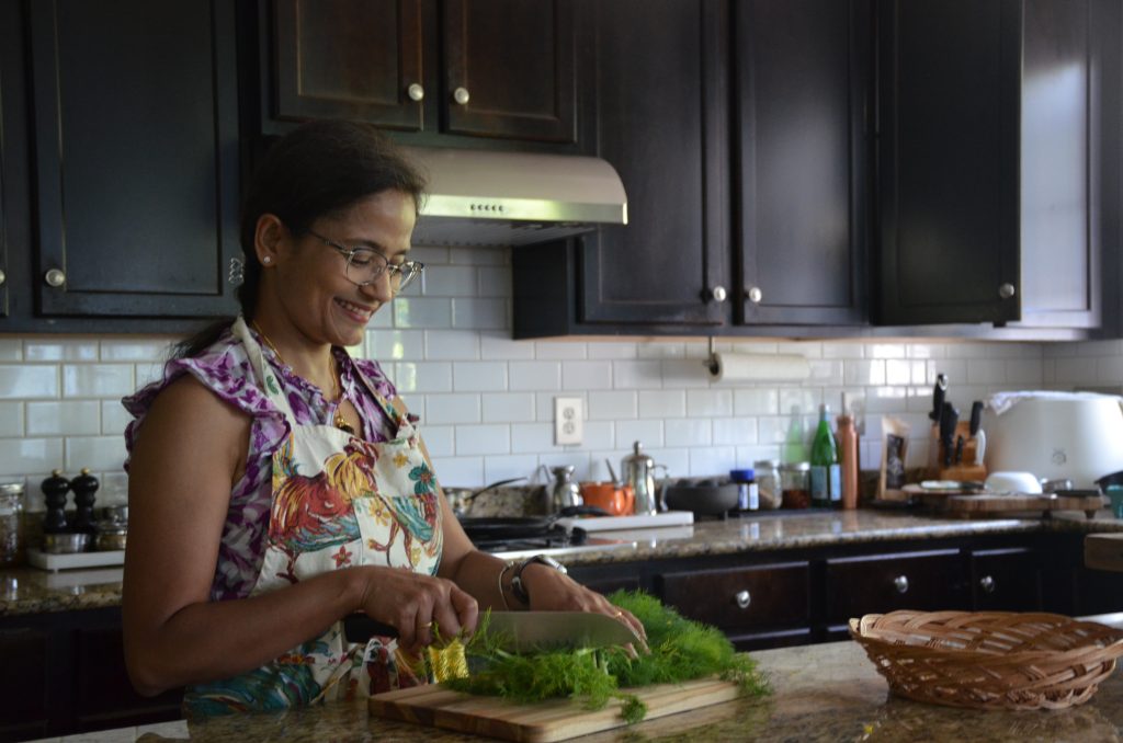 A woman wearing glasses and a floral apron smiles while chopping herbs on a wooden cutting board in a York PA kitchen—reflecting the spirit of Heidelberger Integrative holistic care, surrounded by dark cabinets and granite countertops.