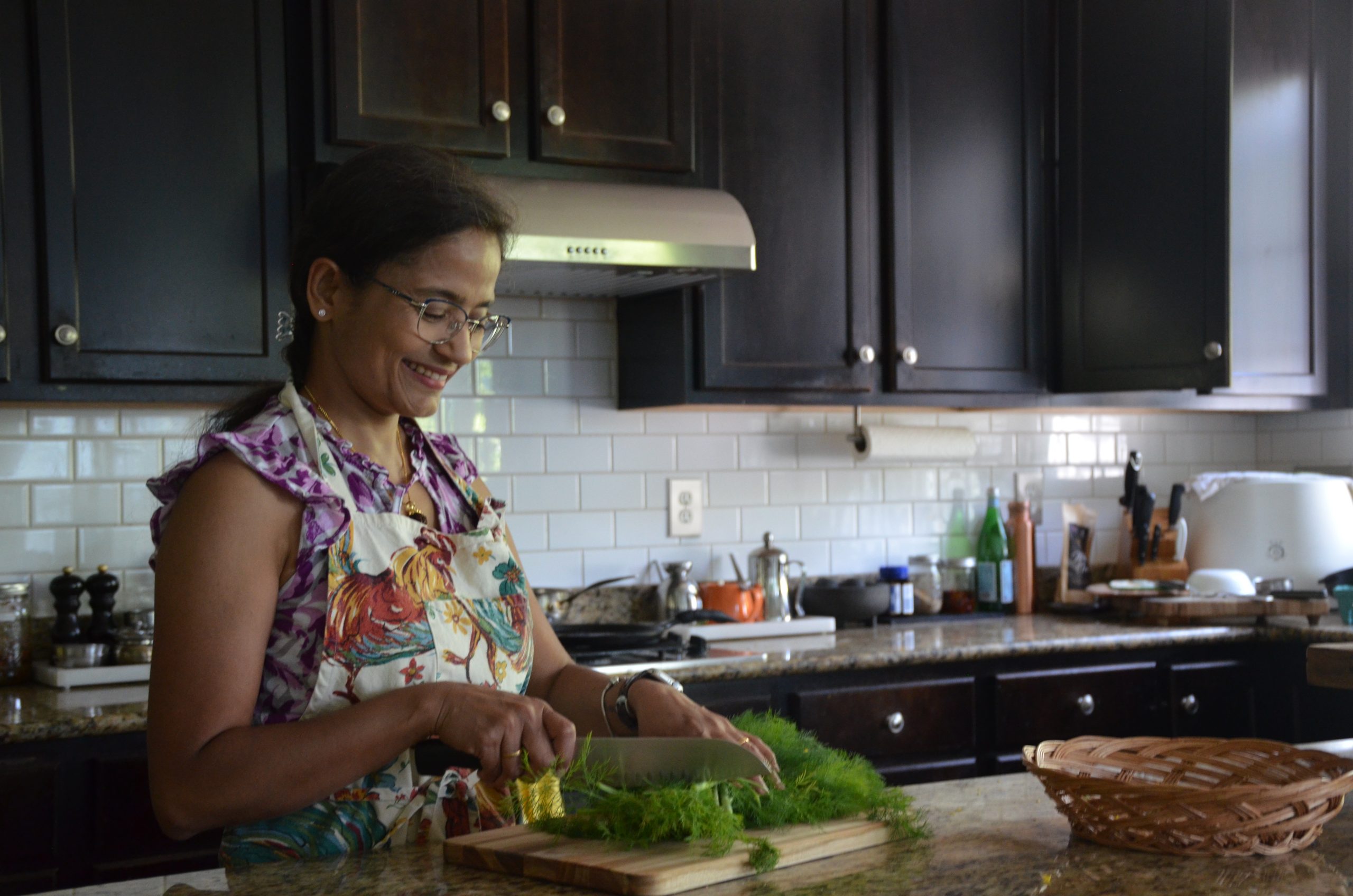 A woman wearing glasses and a floral apron smiles while chopping herbs on a wooden cutting board in a York PA kitchen—reflecting the spirit of Heidelberger Integrative holistic care, surrounded by dark cabinets and granite countertops.