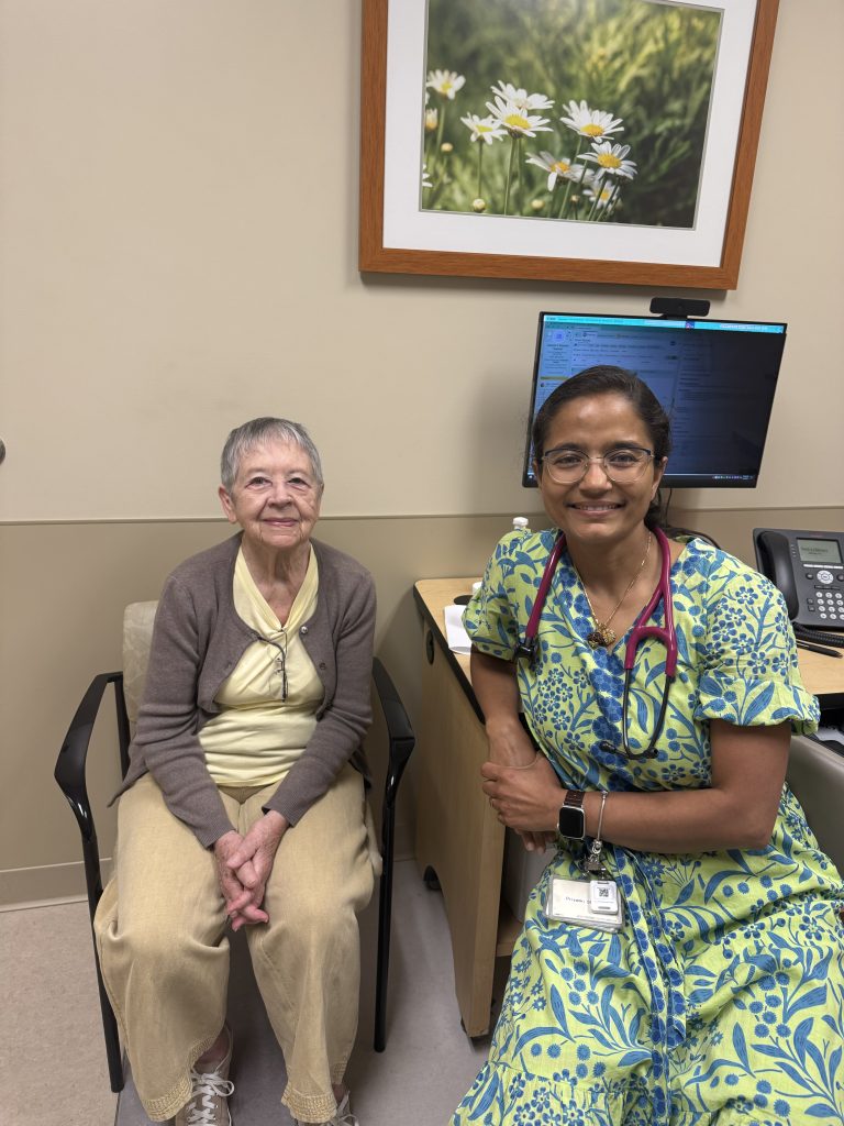 An elderly woman sits beside a smiling doctor in a brightly patterned dress at Heidelberger Integrative in York PA, inside a medical office with a computer, phone, and framed flower photograph—reflecting their commitment to holistic care.