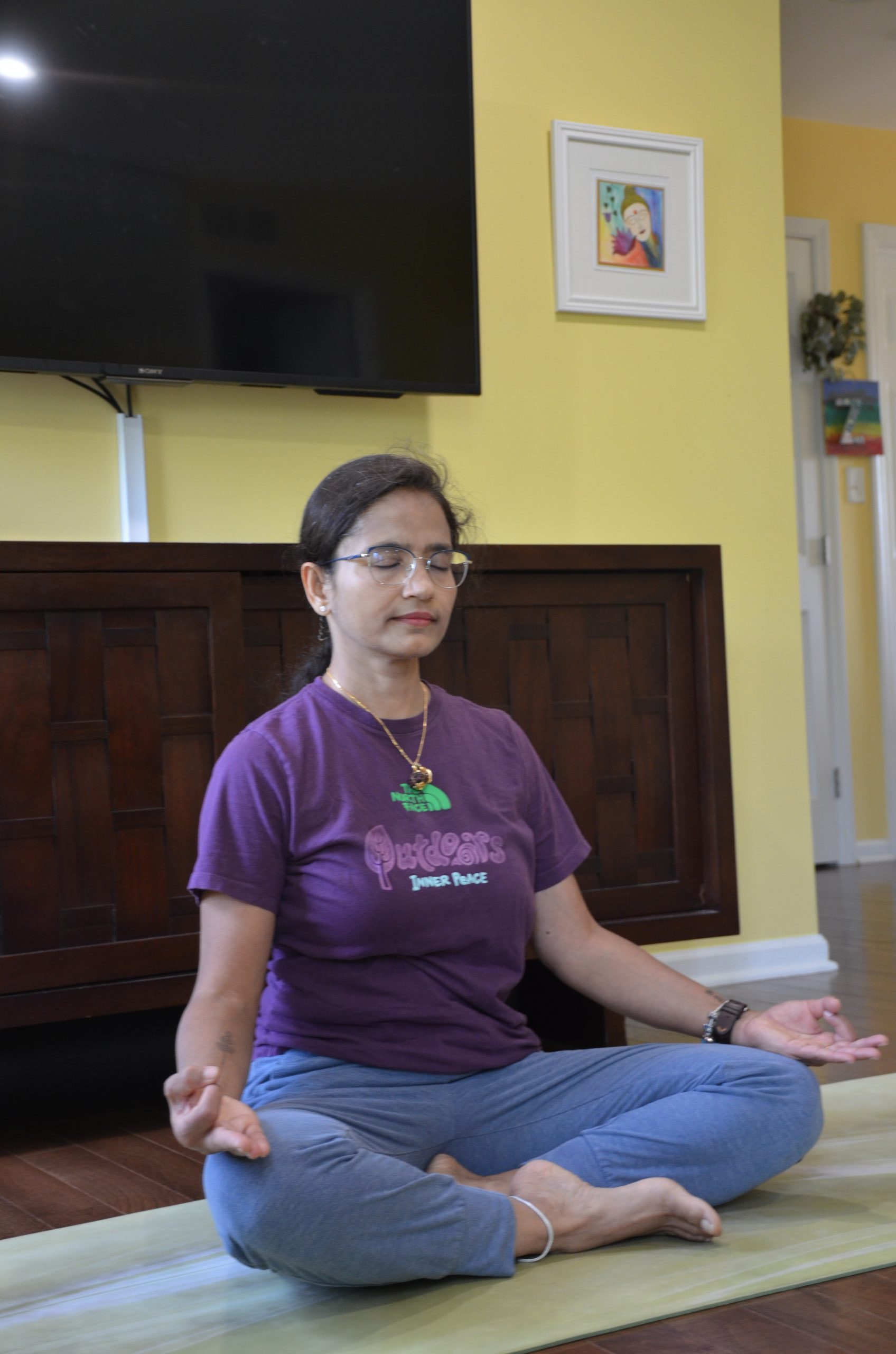 A woman sits cross-legged on a mat indoors, meditating with her eyes closed and embracing wellness. She wears glasses, a purple t-shirt, and blue pants. A TV and framed artwork decorate the yellow wall behind her in York PA.