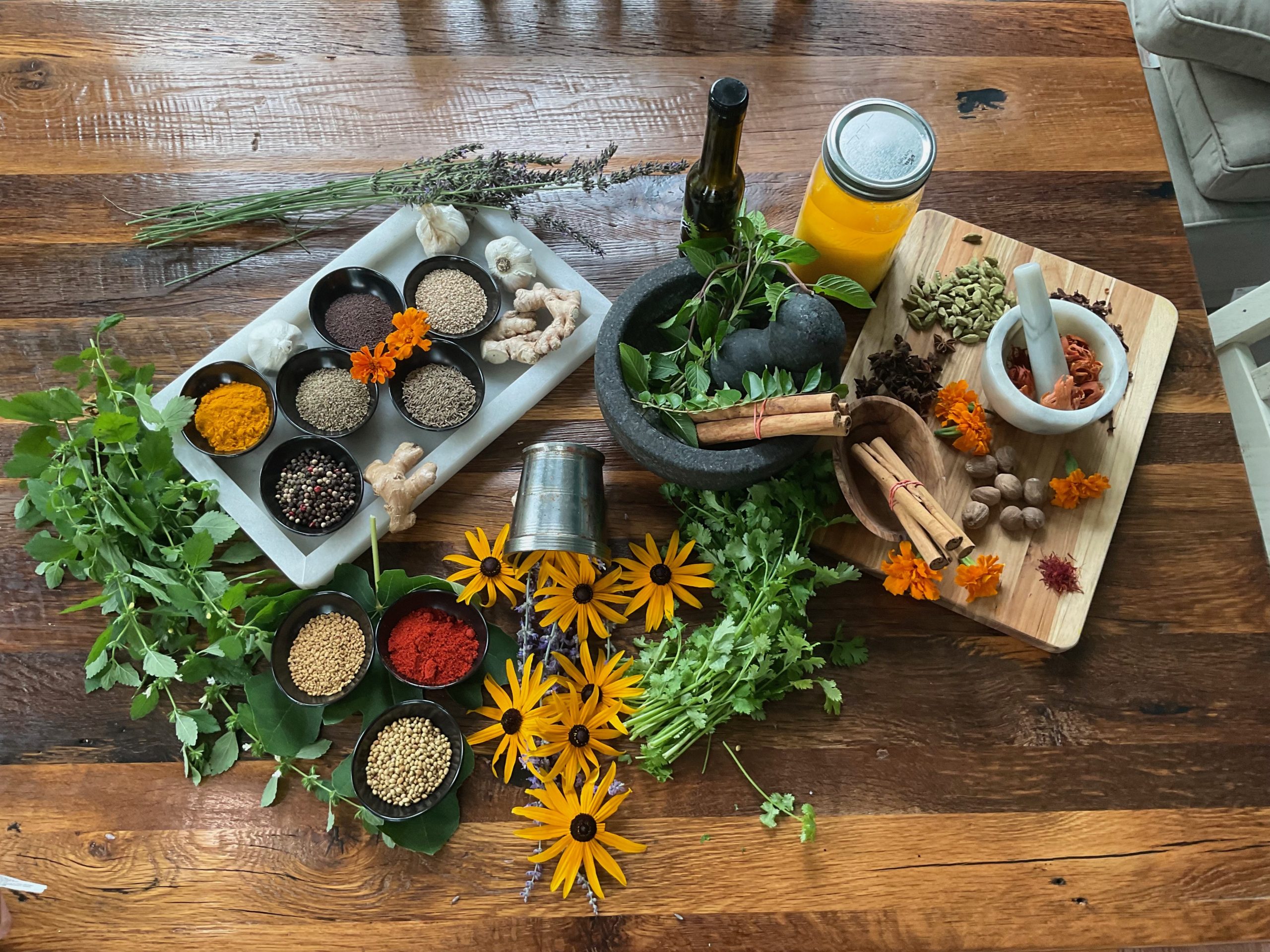 A wooden table displays fresh herbs, flowers, bowls of spices, a mortar and pestle with spices, ginger, a jar of yellow liquid, and a bottle—creating a colorful kitchen scene that reflects Heidelberger Integrative wellness and natural living.