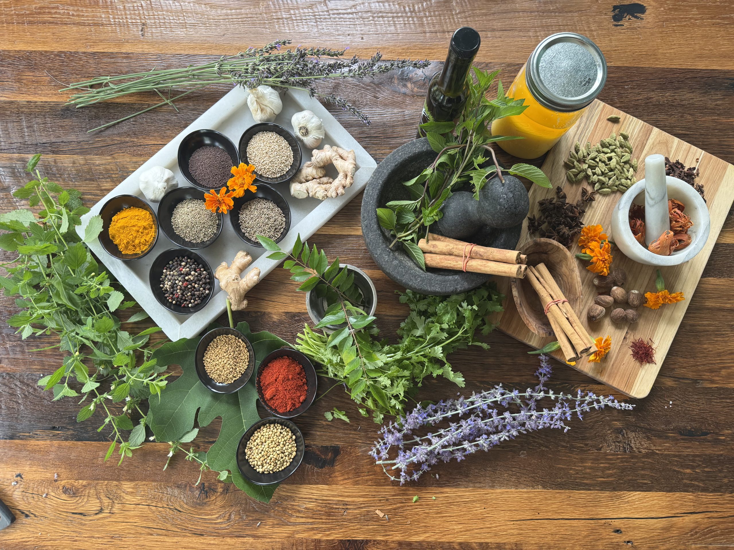 A variety of fresh herbs, spices, and powders for wellness, including turmeric, ginger, cinnamon, and cardamom, are arranged on a wooden table with mortar and pestle, oil, garlic, and flowers—perfect for holistic care in York PA.