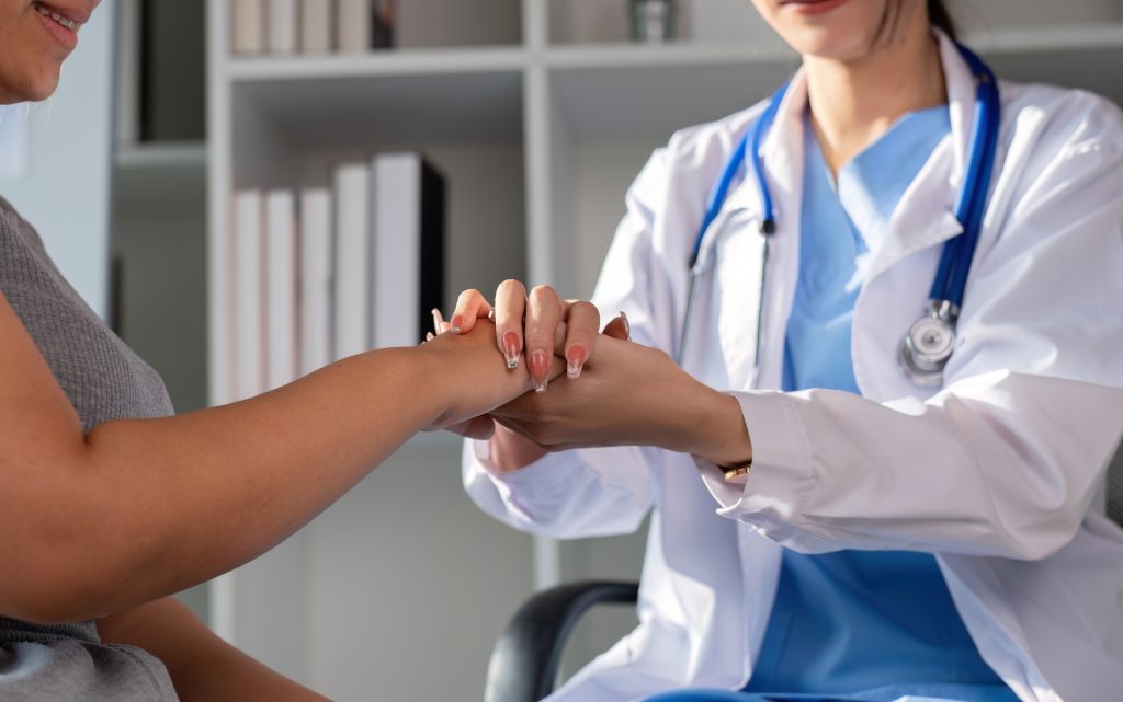 A doctor in a white coat holds and examines a patient’s hand in a medical office, showing care and support. Shelves with books are visible in the background, reflecting a commitment to wellness and integrative medicine.