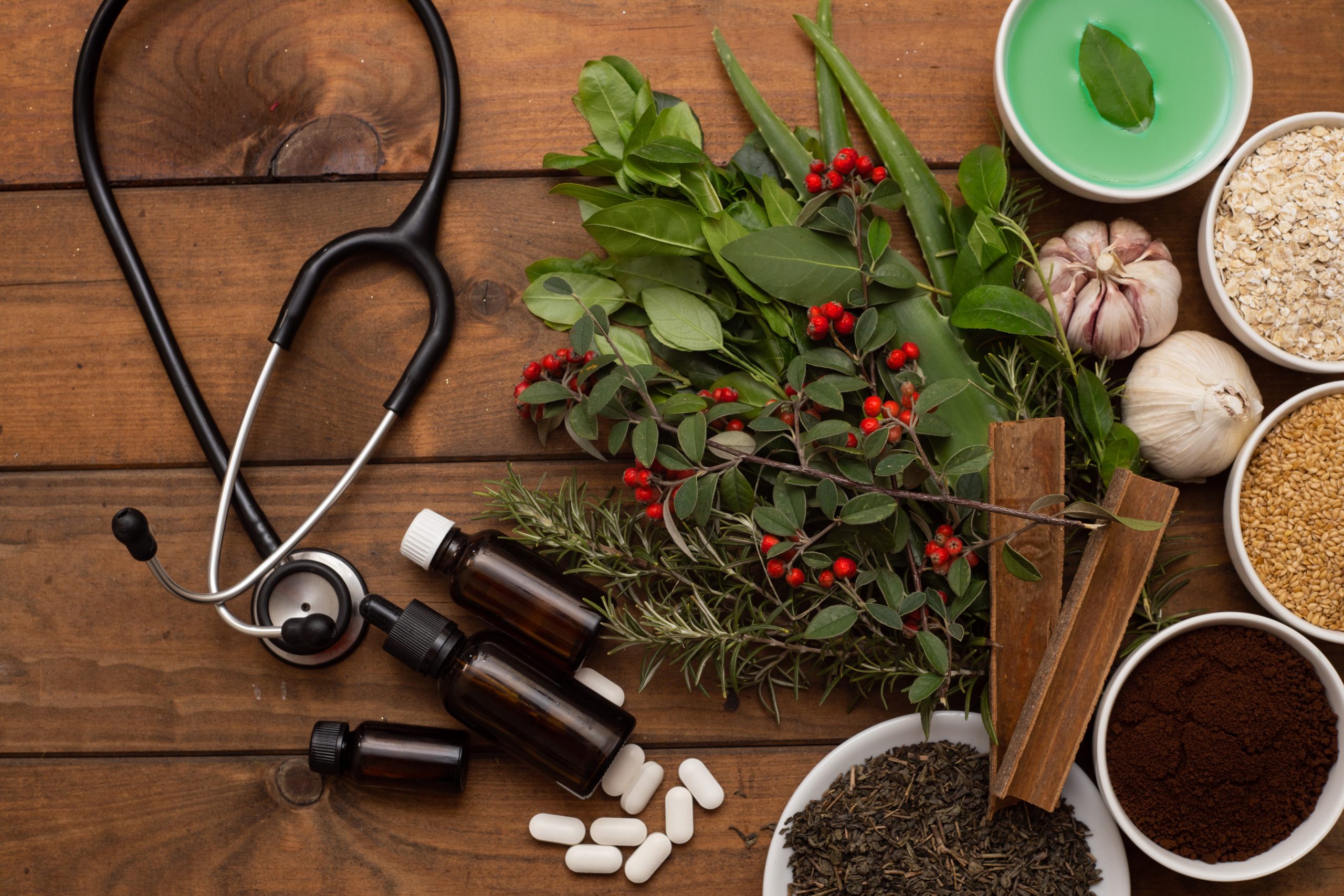 A stethoscope, herbal leaves, garlic, spices, pills, and bowls of seeds and tea are arranged on a wooden table, symbolizing integrative medicine that blends natural remedies with modern healthcare and nutritional guidance.